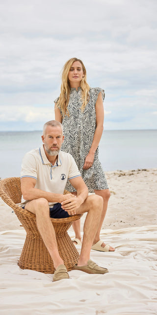 Her ses et afslappet strandlook fra Redgreen – mand i klassisk polo og shorts, og kvinde i let, mønstret kjole med korte ærmer. Perfekt til en rolig dag ved vandet med stil og komfort.