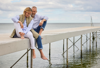 Mand og kvinde i afslappet Redgreen forårsoutfit sidder på en badebro – kvinde i quiltet vest, lyseblå skjorte og hvide bukser, mand i lyseblå skjorte og jeans. Perfekt til hygge ved vandet og tidløst skandinavisk design.